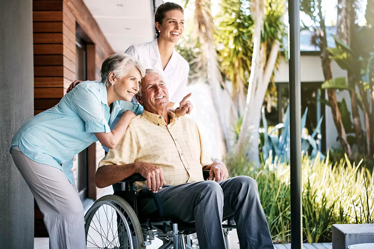 Ein Rollstuhlfahrer schaut zusammen mit seiner Ehefrau und einer Pflegerin in den Innenhof einer Tagespflege.